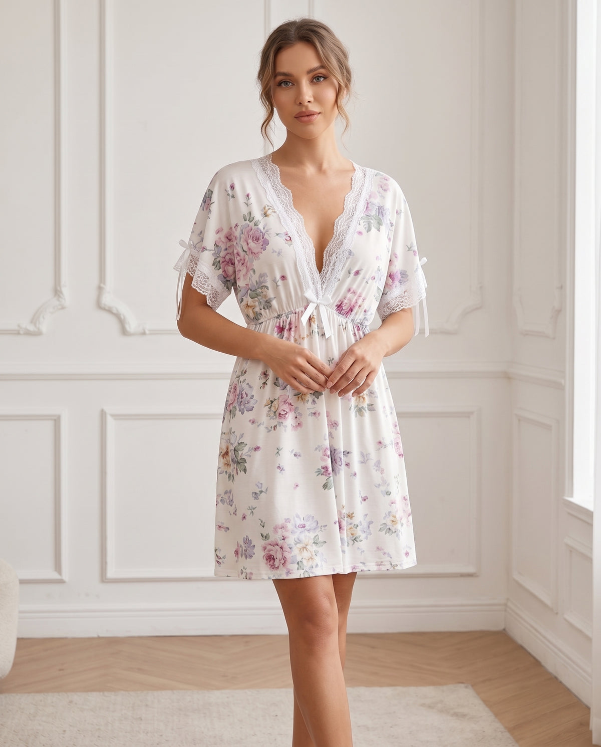 A woman models a short white nightgown with a pastel floral print, lace trim, and a tie waist, standing in a bright room with white paneled walls.
