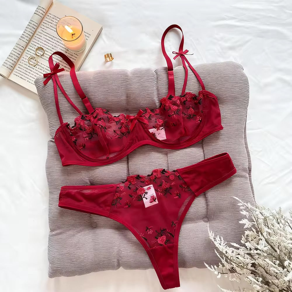 Red lingerie set with floral embroidery on a gray pillow, surrounded by a book, candle, and perfume bottle.