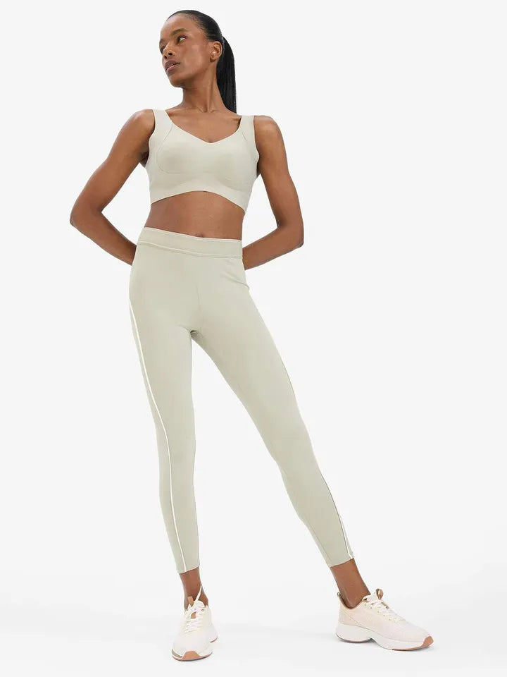 A woman modeling a matching athletic set consisting of a light beige sports bra and high-waisted leggings with white side stripes, paired with white sneakers, against a solid light gray background.