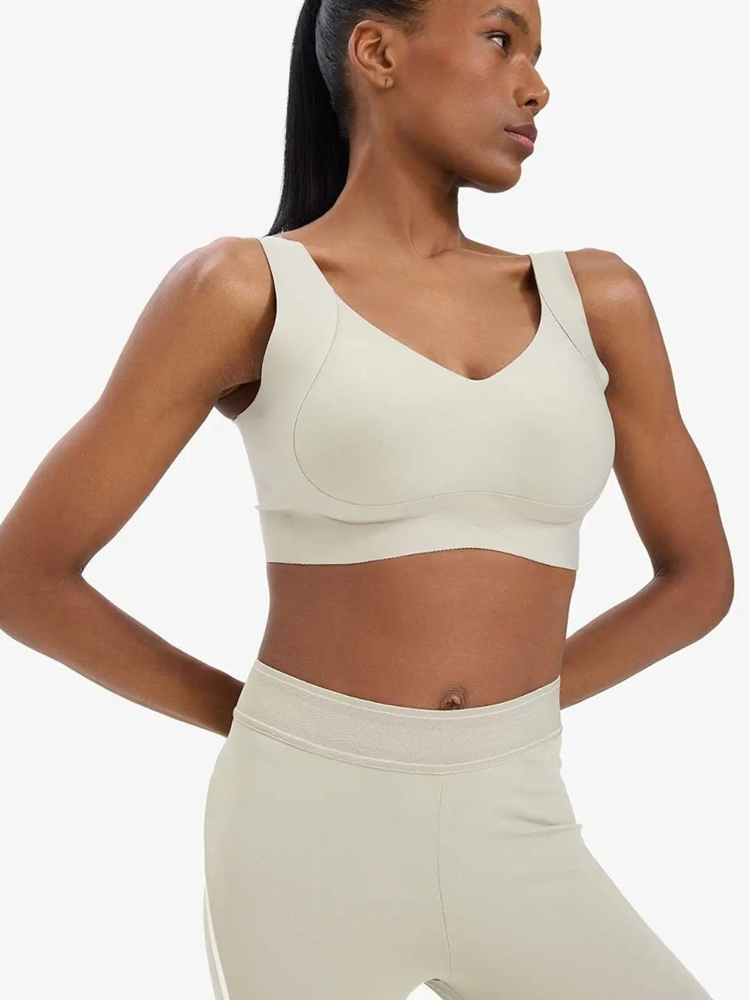 A woman wearing a light beige sports bra and matching high-waisted leggings, shown from the side against a plain light gray studio background.