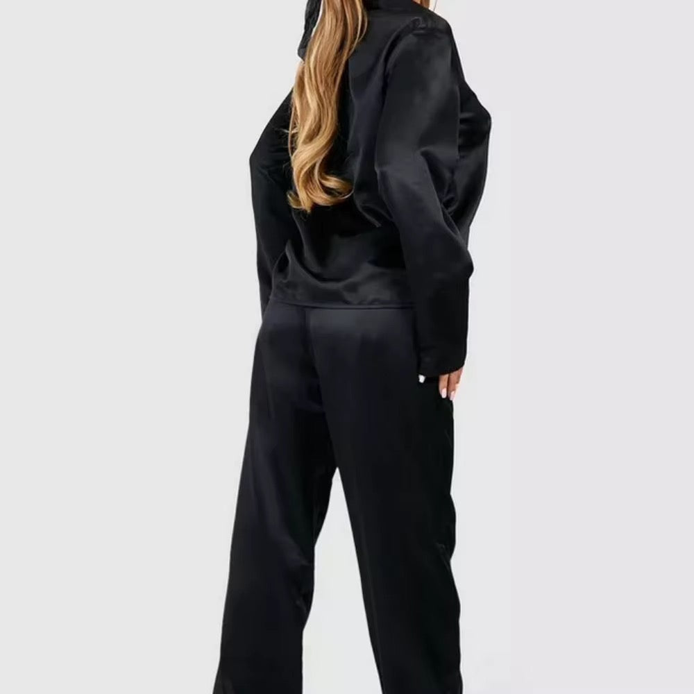 A woman wearing a black satin two-piece tracksuit, viewed from the back, against a plain light gray studio background.