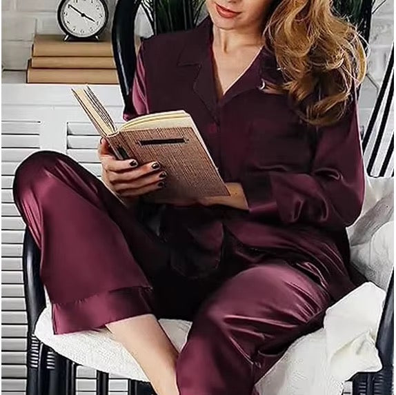 A woman wearing a satin-like burgundy pajama set sits in a chair reading a book. The set includes a long-sleeve button-front top and matching pants, shown in a styled indoor setting with a chair, white slatted wall, books, and a clock in the background.