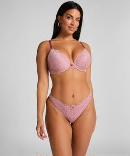 A woman modeling a light pink lace lingerie set against a plain light gray studio background.