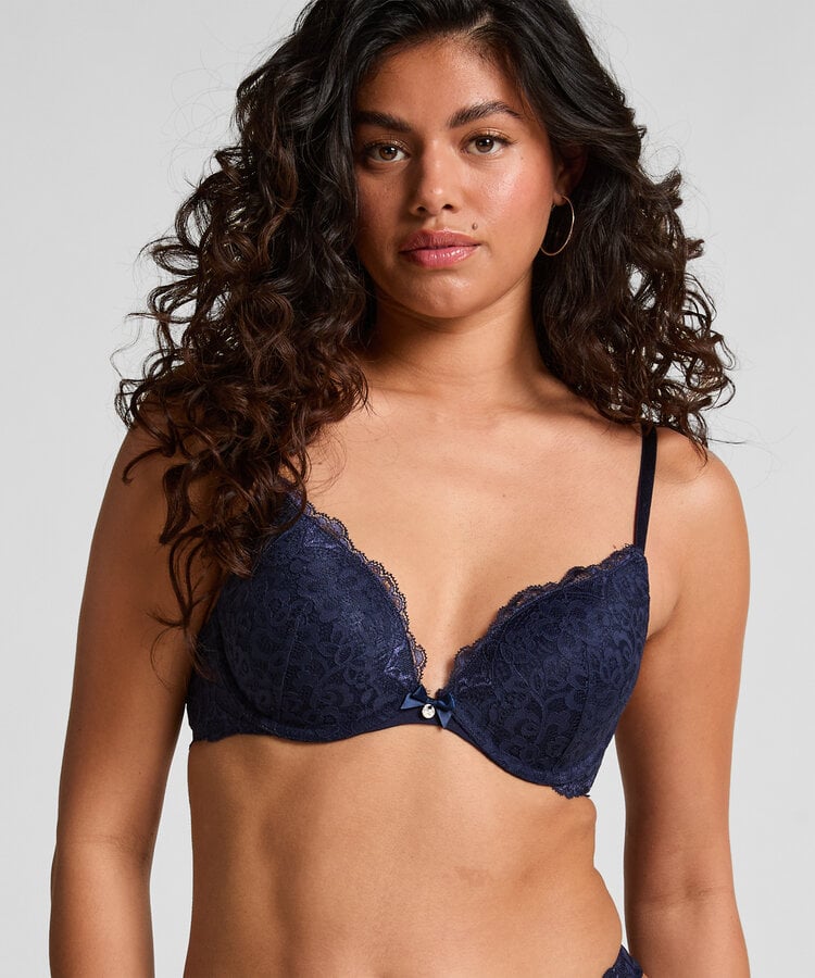 A woman modeling a navy blue lace underwire bra against a light gray studio background.