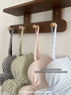 A dark brown wooden wall-mounted rack with four hanging hooks holding striped bras in black-and-white, sage green-and-white, pink-and-white, and light blue-and-white gingham patterns.