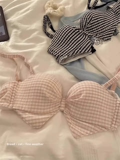 A light pink and white gingham check bikini top laid flat on white bedding, with a matching striped bikini top and a light blue bikini bottom visible in the background.