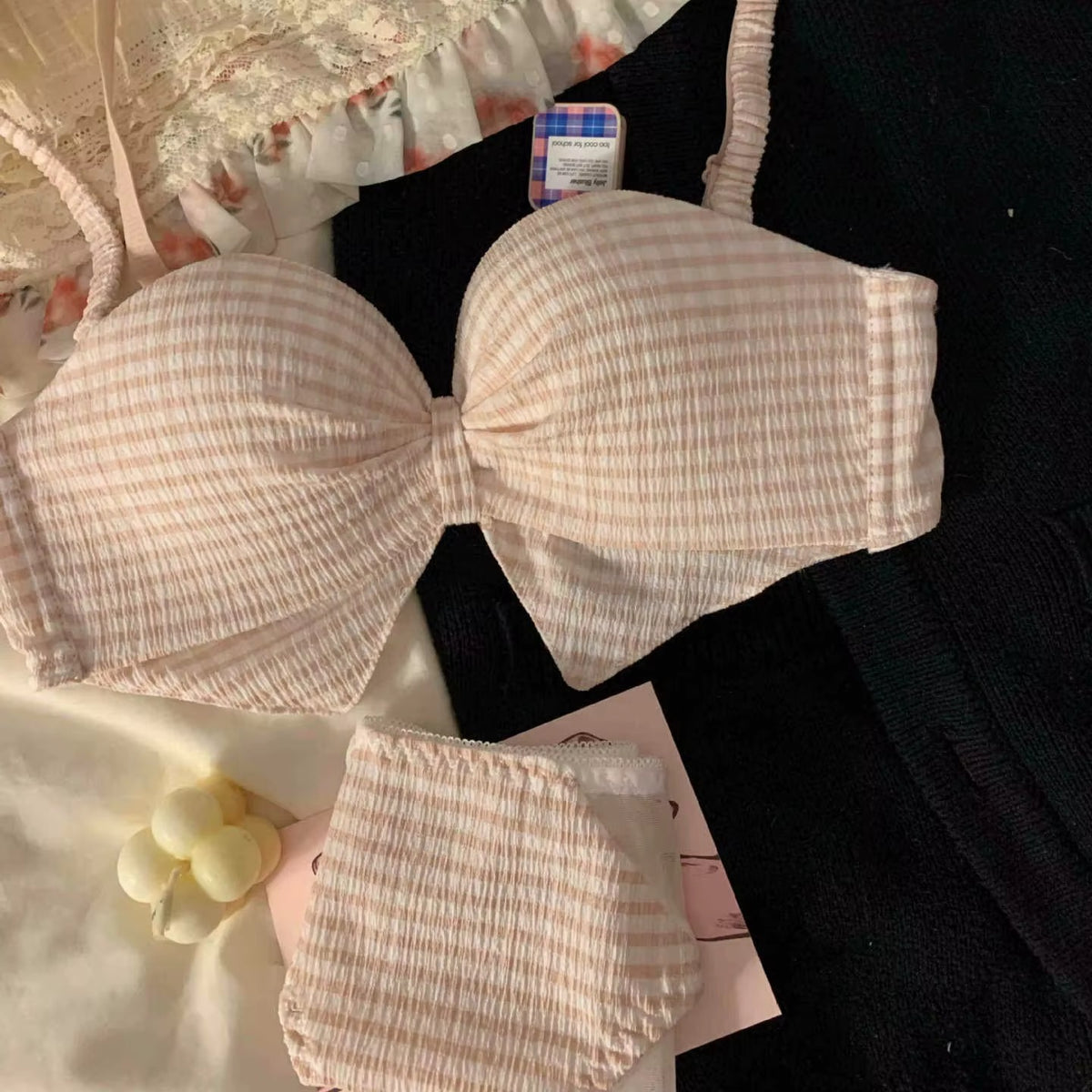 A light pink and white gingham crinkled cotton bralette with a large bow at the center, displayed alongside matching underwear and a small cream-colored flower-shaped candle on a black fabric background.