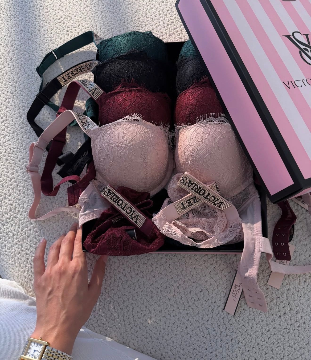 A Victoria's Secret lingerie gift set displayed in a black box on a textured white surface. The set includes multiple lace underwire bras in shades of pink, burgundy, green, and black, along with matching underwear and a pink striped Victoria's Secret box.