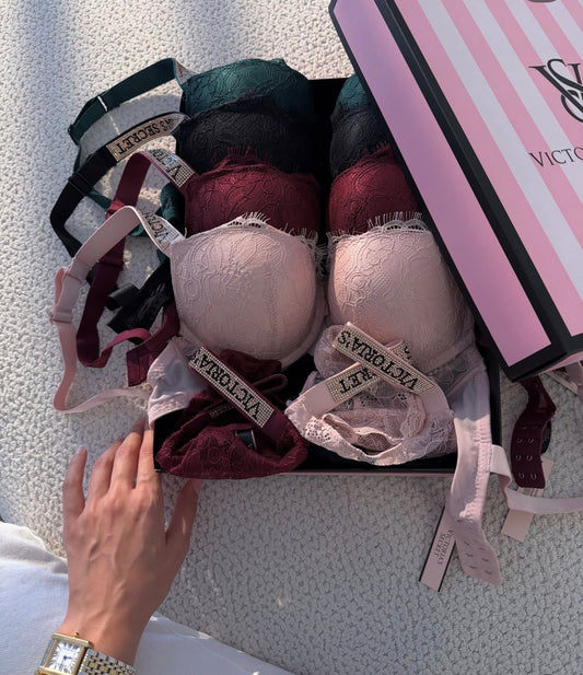 A Victoria's Secret lingerie gift set displayed in a black box on a textured white surface. The set includes multiple lace underwire bras in shades of pink, burgundy, green, and black, along with matching underwear and a pink striped Victoria's Secret box.
