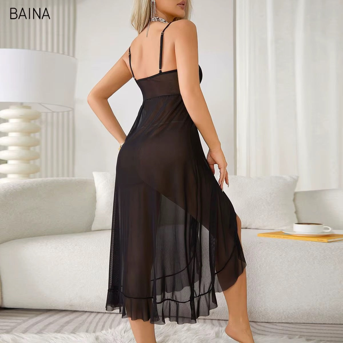 A woman modeling a sheer black midi slip dress with thin spaghetti straps and a backless open back, shown in a bright living room setting. The brand name BAINA is visible in the top left corner.