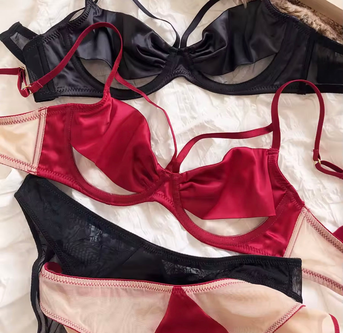 Three satin lingerie sets arranged on a white fabric background: a black bra at the top, a red and beige bra in the center, and a matching black and red panty at the bottom.