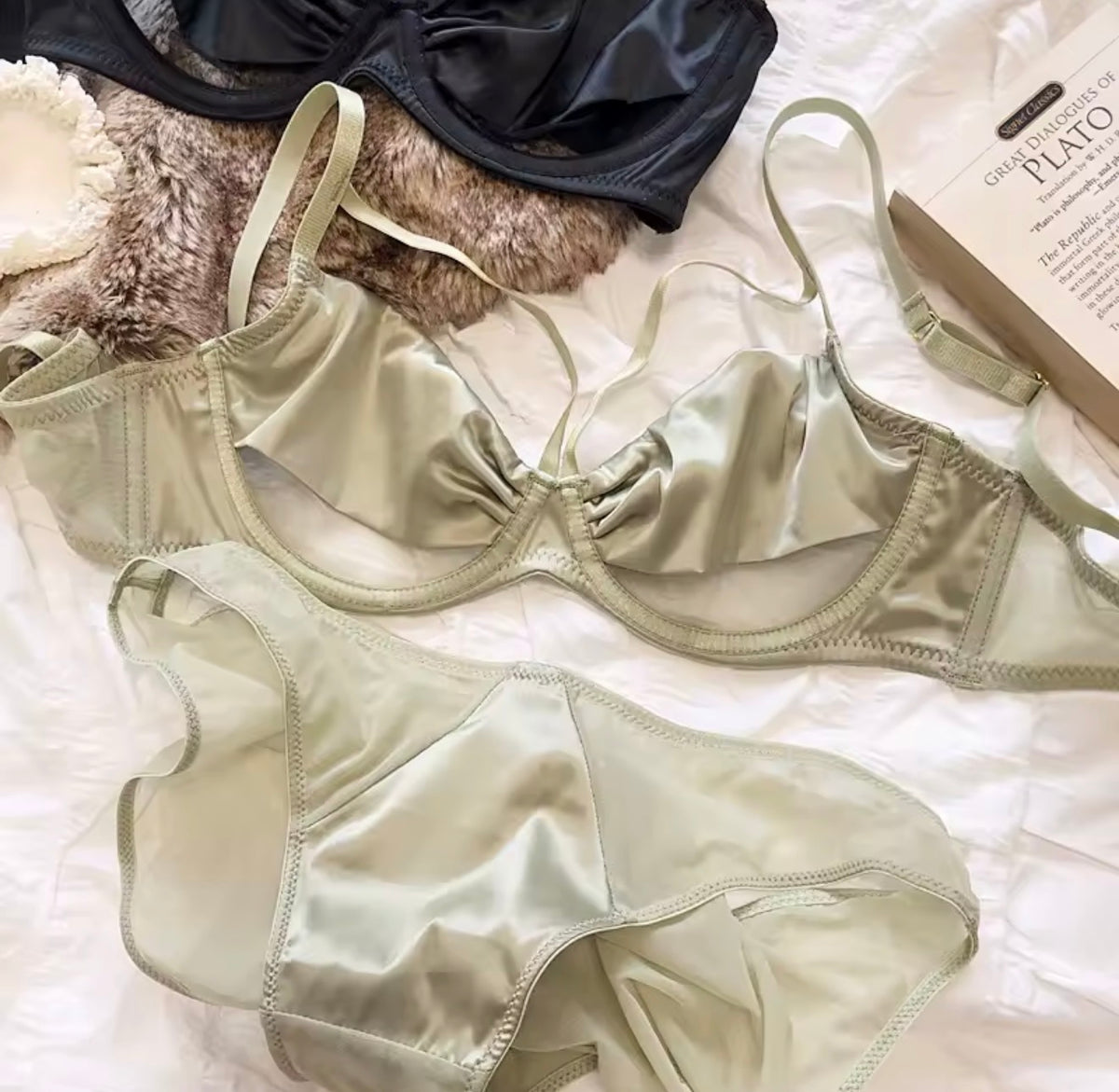 A sage green satin bra and matching underwear set laid flat on a white textured fabric, with a black bra and a book visible as styling props.