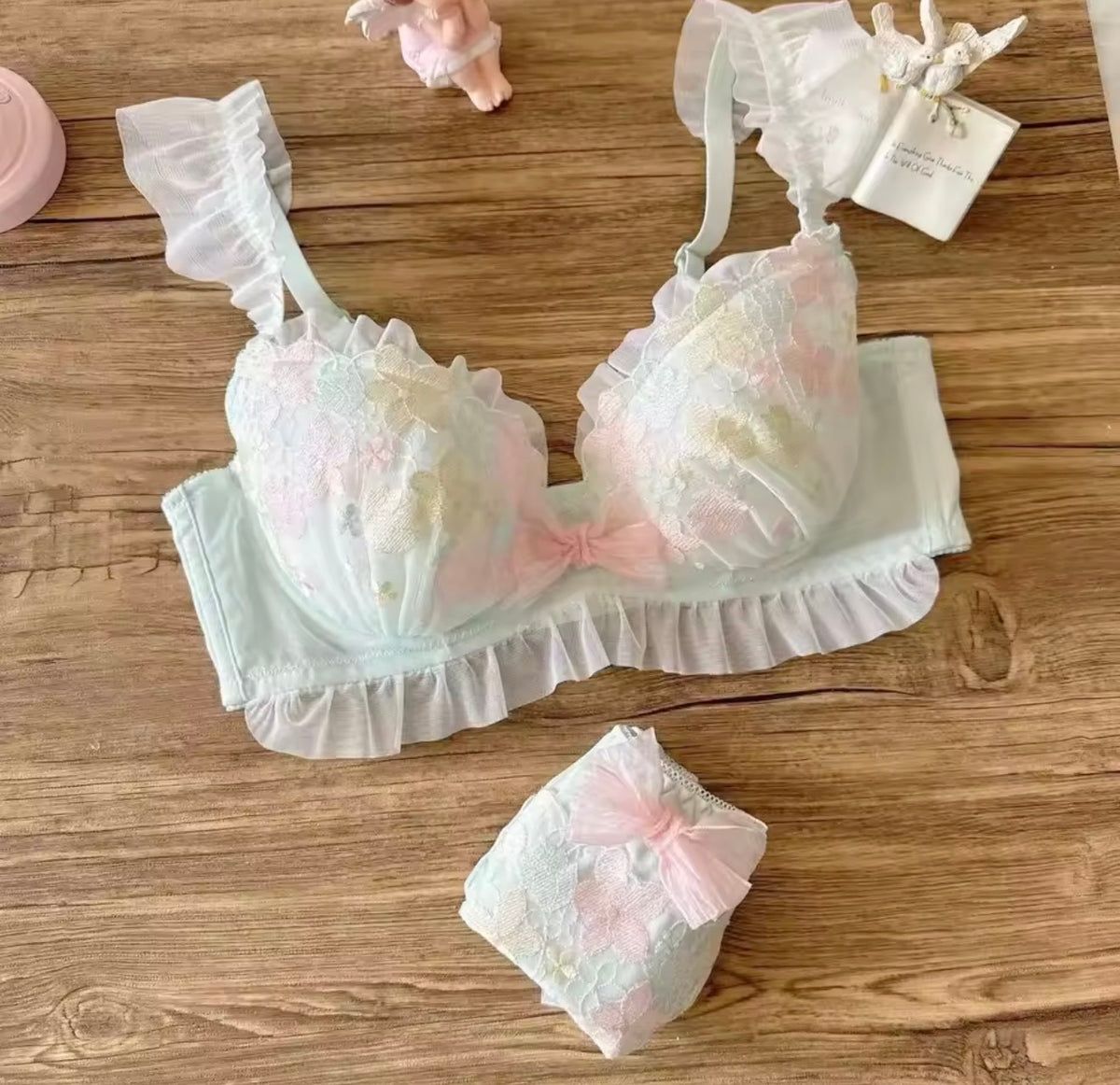 A pastel lace lingerie set laid flat on a wooden surface, including a ruffled bra with soft pink and cream floral detailing, matching panties, and a small decorative tag nearby.