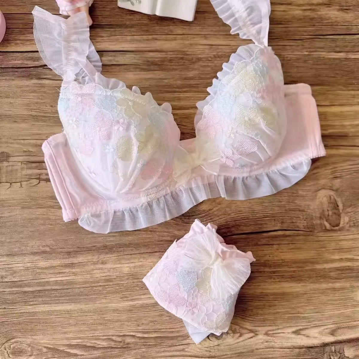 A pastel pink lace bra with ruffled straps and trim, shown laid flat on a wooden surface with matching panties below.