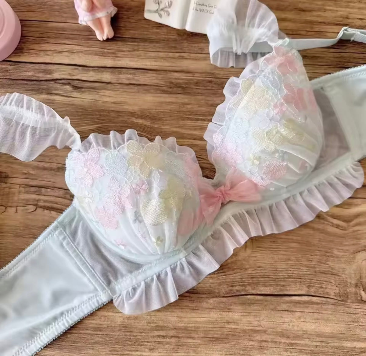 A pastel lace bra with ruffled trim and floral applique in soft pink, white, and pale yellow, laid flat on a wooden surface with matching underwear pieces nearby.