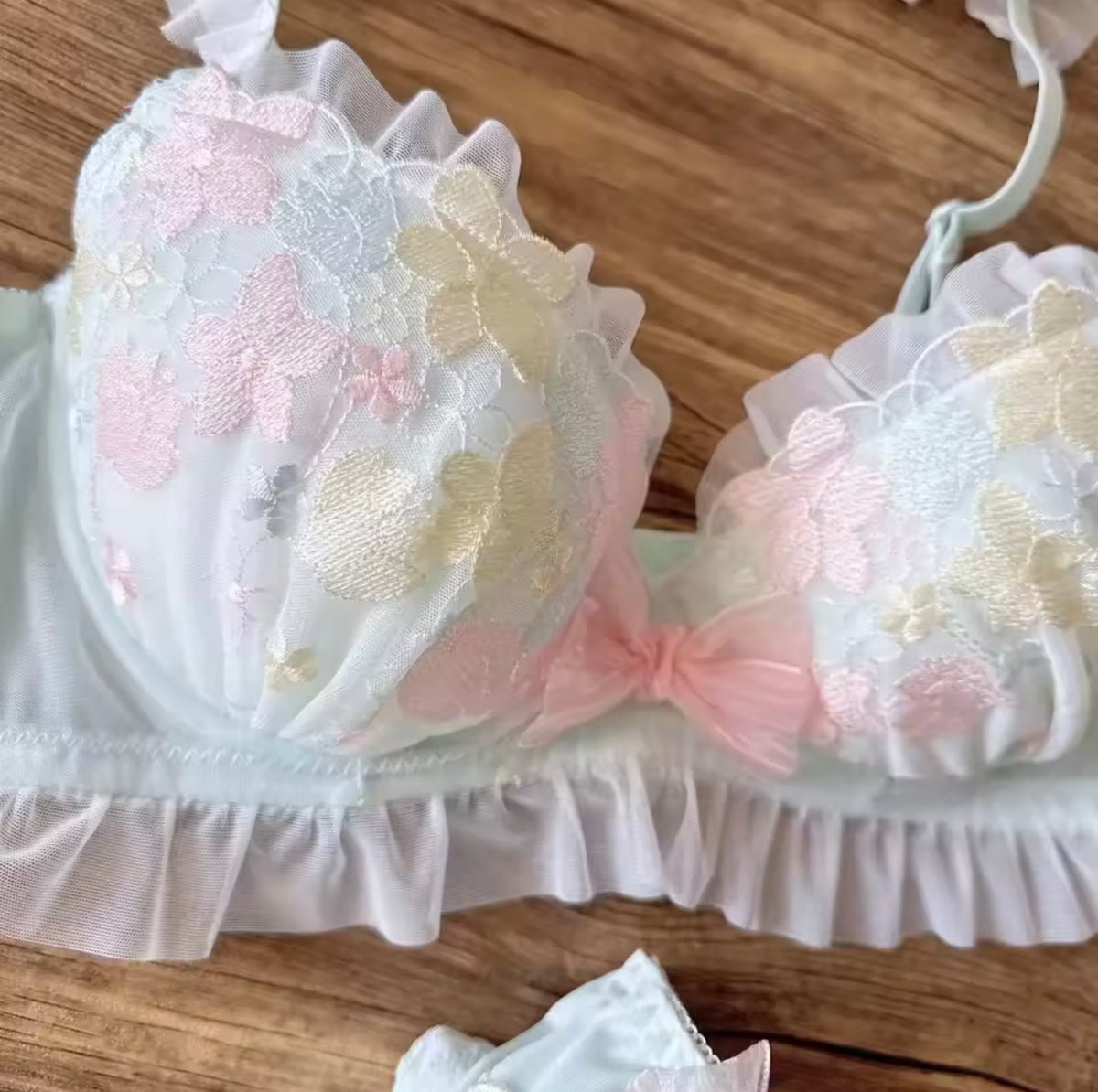 A white ruffled lace bra with pastel pink, yellow, and blue embroidered flowers, displayed on a wooden surface.