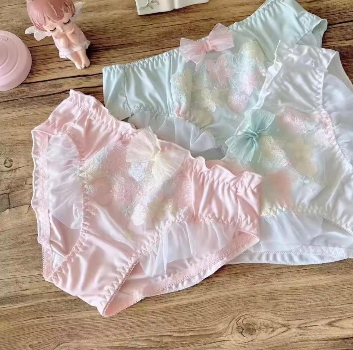 Two pairs of pastel lace panties laid flat on a wooden surface: one light pink with a bow and floral lace, and one white with a pale mint bow and floral lace. Small decorative figurines and a pink container are partially visible in the background.