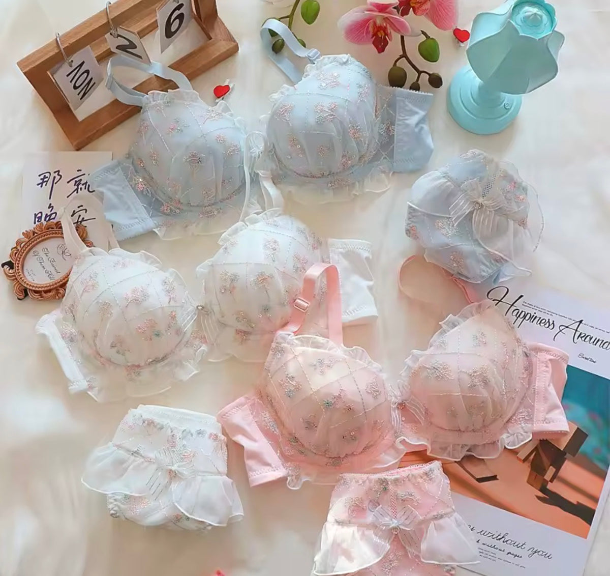 A styled flat-lay of multiple lace bra and matching panty sets in soft pastel colors, including white, pale blue, and pale pink, arranged with decorative props on a light fabric background.