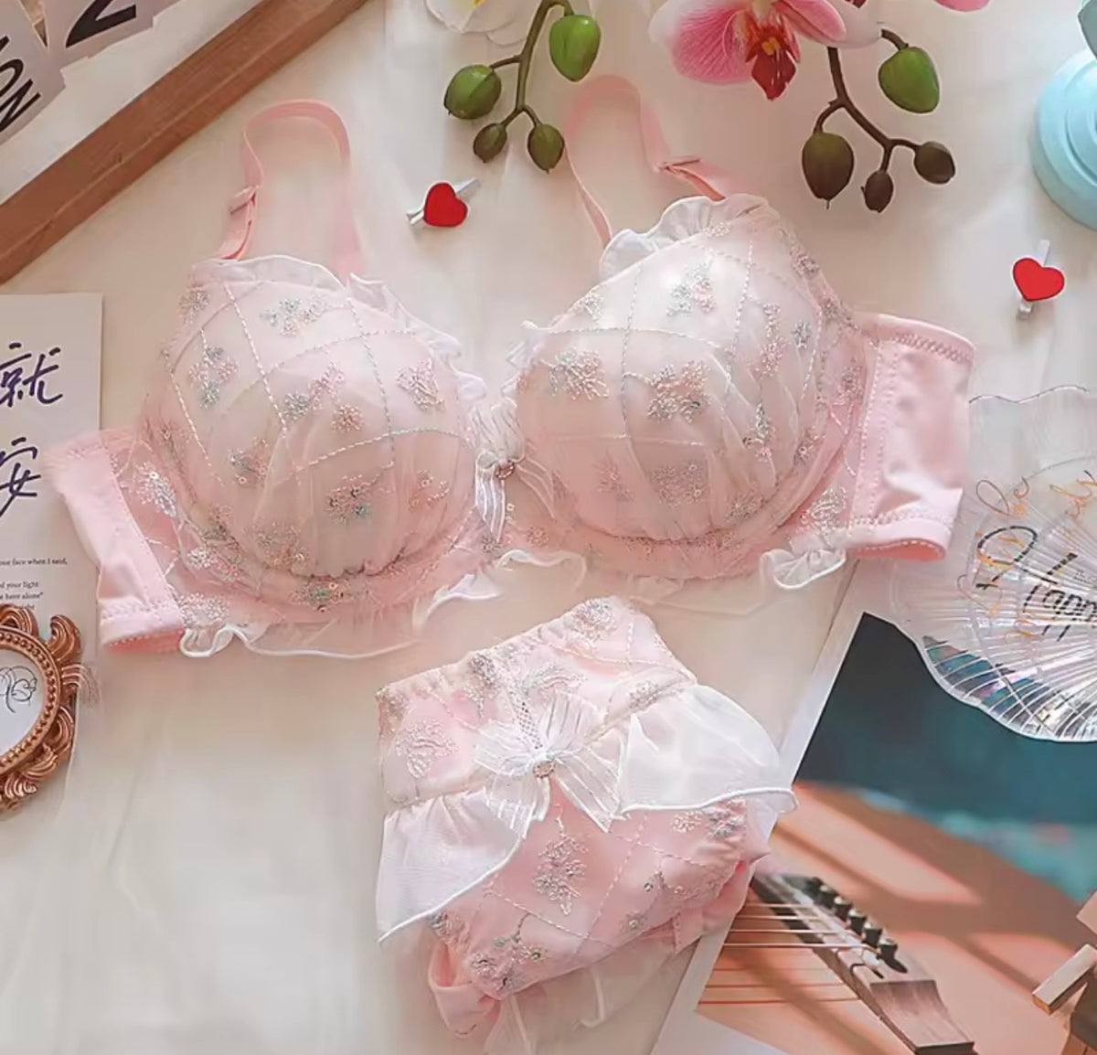 A pink lingerie set laid flat on a light surface, including a sheer floral underwire bra with ruffled trim and matching panties with a large bow. Decorative flowers, small hearts, and printed cards surround the product.