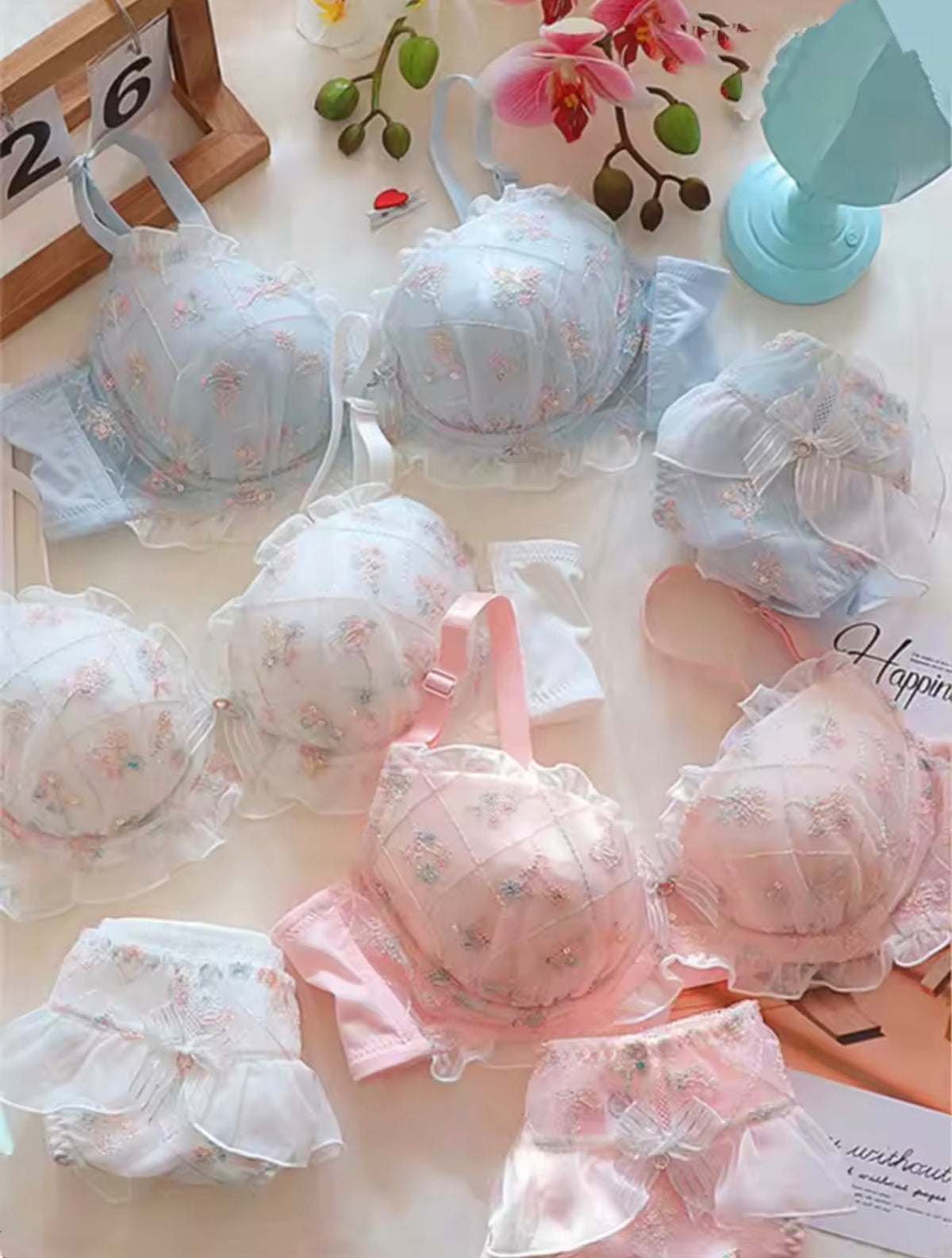 A styled flat-lay display of multiple lace-trimmed underwire bras and matching panties in pastel shades of light blue, pink, and white, decorated with delicate floral prints and ruffled trim.
