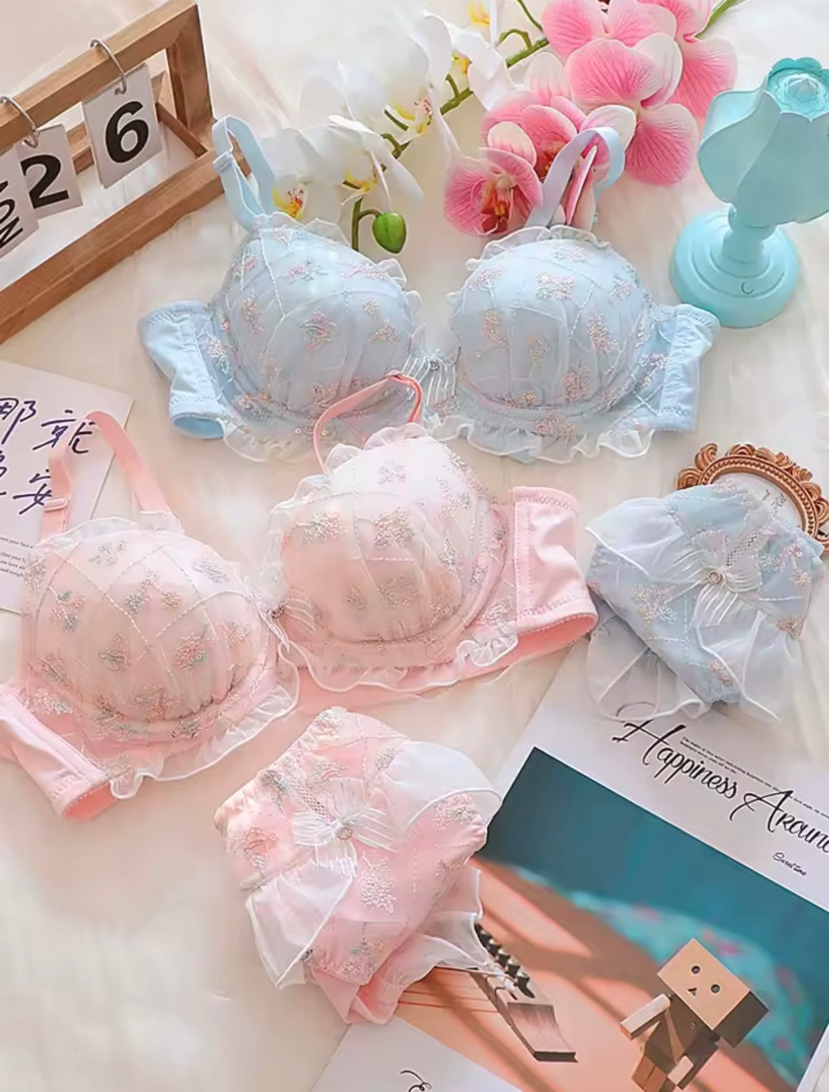 Four pairs of women's lace bra and matching panty set in pastel shades of light blue and pink, decorated with delicate floral prints. The sets are styled in a flat-lay arrangement with decorative props including pink orchids, a light blue lamp, a wooden calendar, and a magazine on a soft beige fabric background.