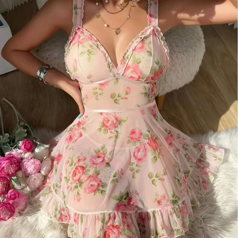 A woman wearing a light pink sheer mesh mini dress with a pink and green rose floral print, lace trim, and ruffled details. She is sitting on a white faux fur chair in a room with wooden floors, with a bouquet of pink roses on the side.