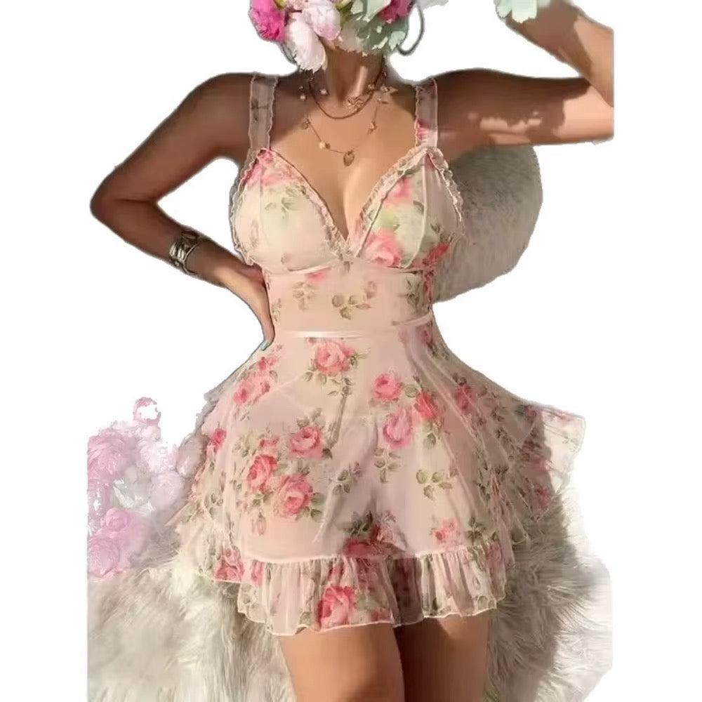 A woman models a sheer pale pink lingerie romper with a pink and green rose floral print, featuring lace trim on the cups and a ruffled hem, styled with a fluffy white faux-fur accessory against a plain white background.