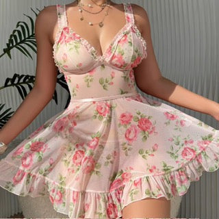 A woman wearing a light pink floral romper dress with a ruffled skirt, featuring a pink rose pattern with green leaves. The image is cropped to show the model from the shoulders down, with a leafy plant visible on the left side against a gray paneled wall.