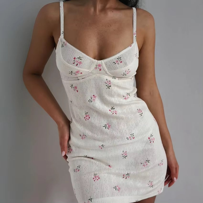 A woman wearing a white ribbed spaghetti-strap mini dress with a small pink and green floral print, photographed against a plain gray wall.