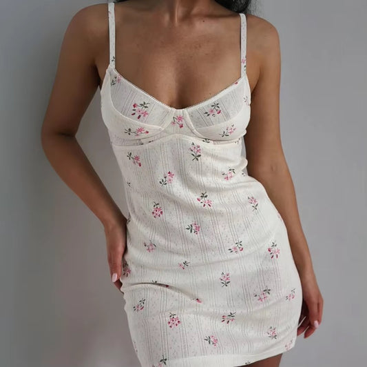 A woman wearing a white ribbed spaghetti-strap mini dress with a small pink and green floral print, photographed against a plain gray wall.