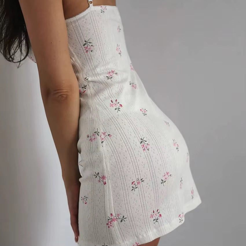 A woman wearing a white textured mini dress with a delicate pink and green floral print, shown from a side-back angle against a plain light gray background.