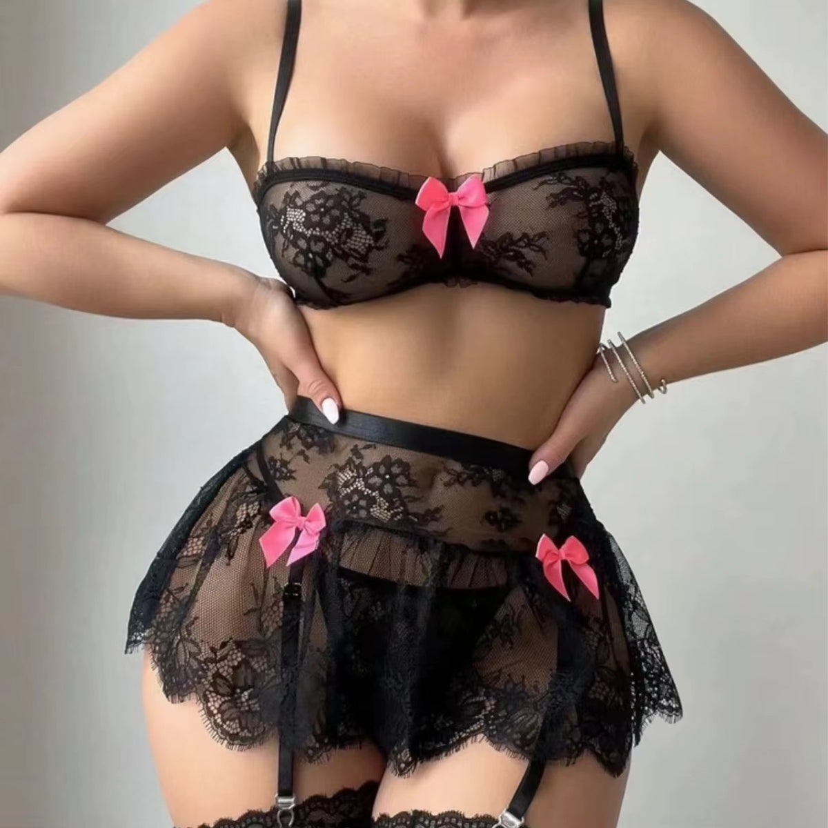 A model wearing a black lace lingerie set with pink bow accents, featuring a sheer bralette, matching mini skirt, and garter belt.
