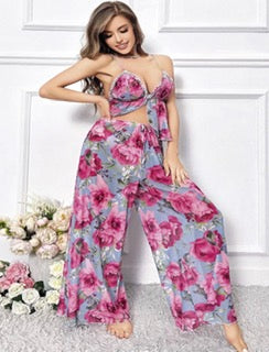 A woman models a two-piece floral jumpsuit set featuring a halter crop top and matching wide-leg pants in a pink and purple floral print on a light blue-gray background.