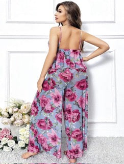A woman modeling a sleeveless wide-leg jumpsuit with a pink and purple floral print on a light blue-gray background, shown from the back against a white paneled wall.