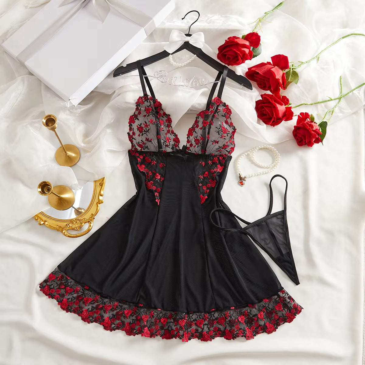 Black lingerie set with red floral lace details, displayed on a hanger with a matching thong, styled on white satin with red roses, gold jewelry, and a white box.