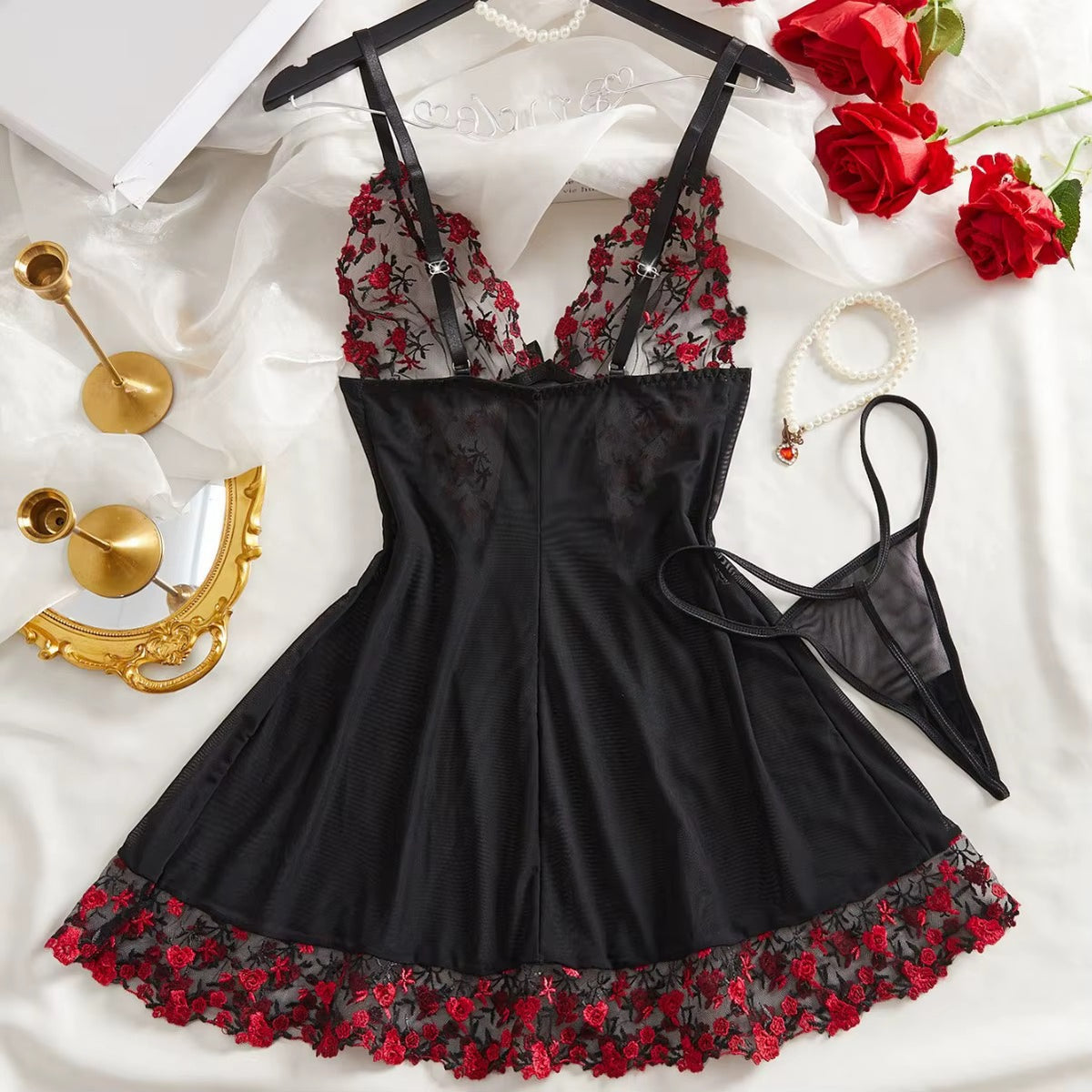 Black lingerie bodysuit with red floral lace cups and hem, displayed on a hanger with a matching thong, styled on white satin with red roses and gold accessories.
