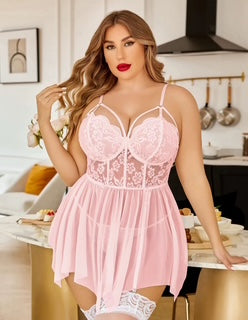 A woman models a light pink lace and mesh babydoll lingerie dress in a bright kitchen setting. The dress features a lace corset-style bodice with thin straps, a sheer mesh skirt, and white lace thigh-high stockings.