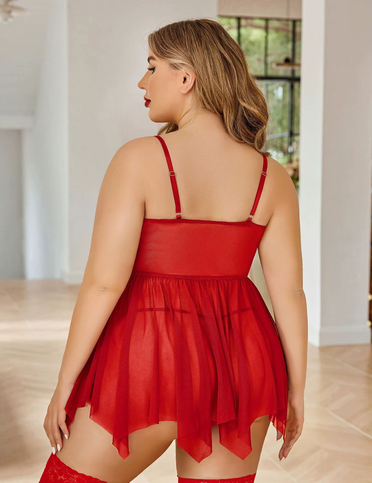 A woman wearing a red sheer babydoll lingerie dress, shown from the back in a bright indoor room with light wood floors and white walls.